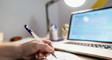 Person füllt Steuererklärung mit Stift auf Papier aus. Im Hintergrund: Laptopbildschirm mit Steuerprogramm.