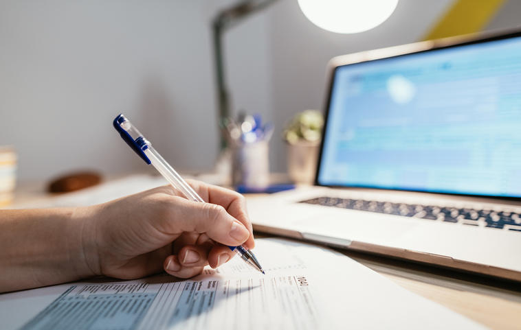 Person füllt Steuererklärung mit Stift auf Papier aus. Im Hintergrund: Laptopbildschirm mit Steuerprogramm.