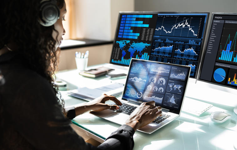 Frau arbeitet an Laptop und hat zwei externe Bildschirme angeschlossen. Alle Displays zeigen Statistiken an.