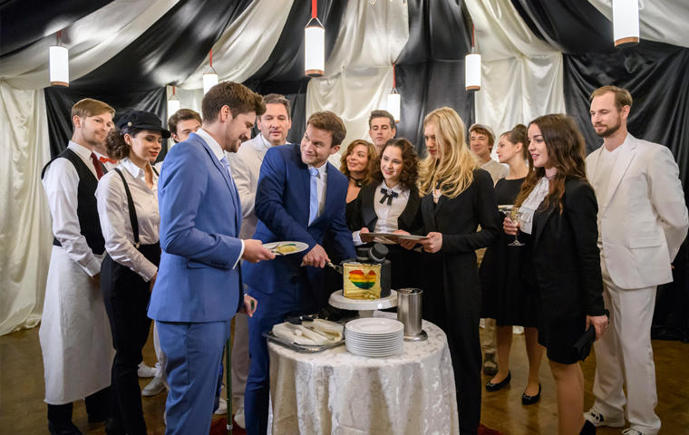 umringt von Hochzeitsgästen schneiden Tobias (Max Beier, l.) und Boris (Florian Frowein, r.) die Hochzeitstorte an, verziert mit einem Herz in Regenbogenfarben.